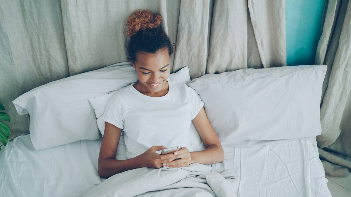 Cheerful African American lady is using smartphone lying in bed under warm blanket on soft pillow. Bedtime, communication and modern technology concept.