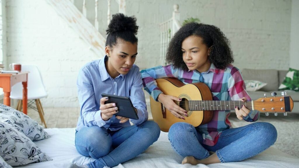 Mixed race young woman with tablet computer sitting on bed teaching her teenage sister to play acoustic guitar