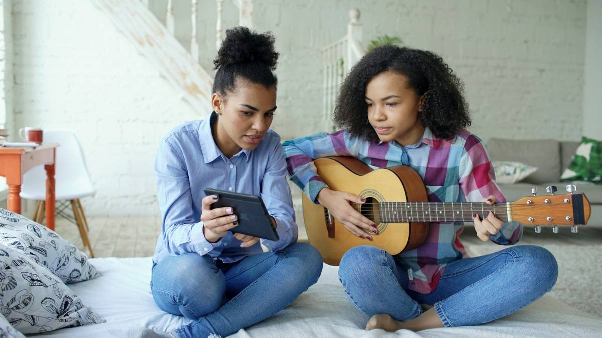 Mixed race young woman with tablet computer sitting on bed teaching her teenage sister to play acoustic guitar